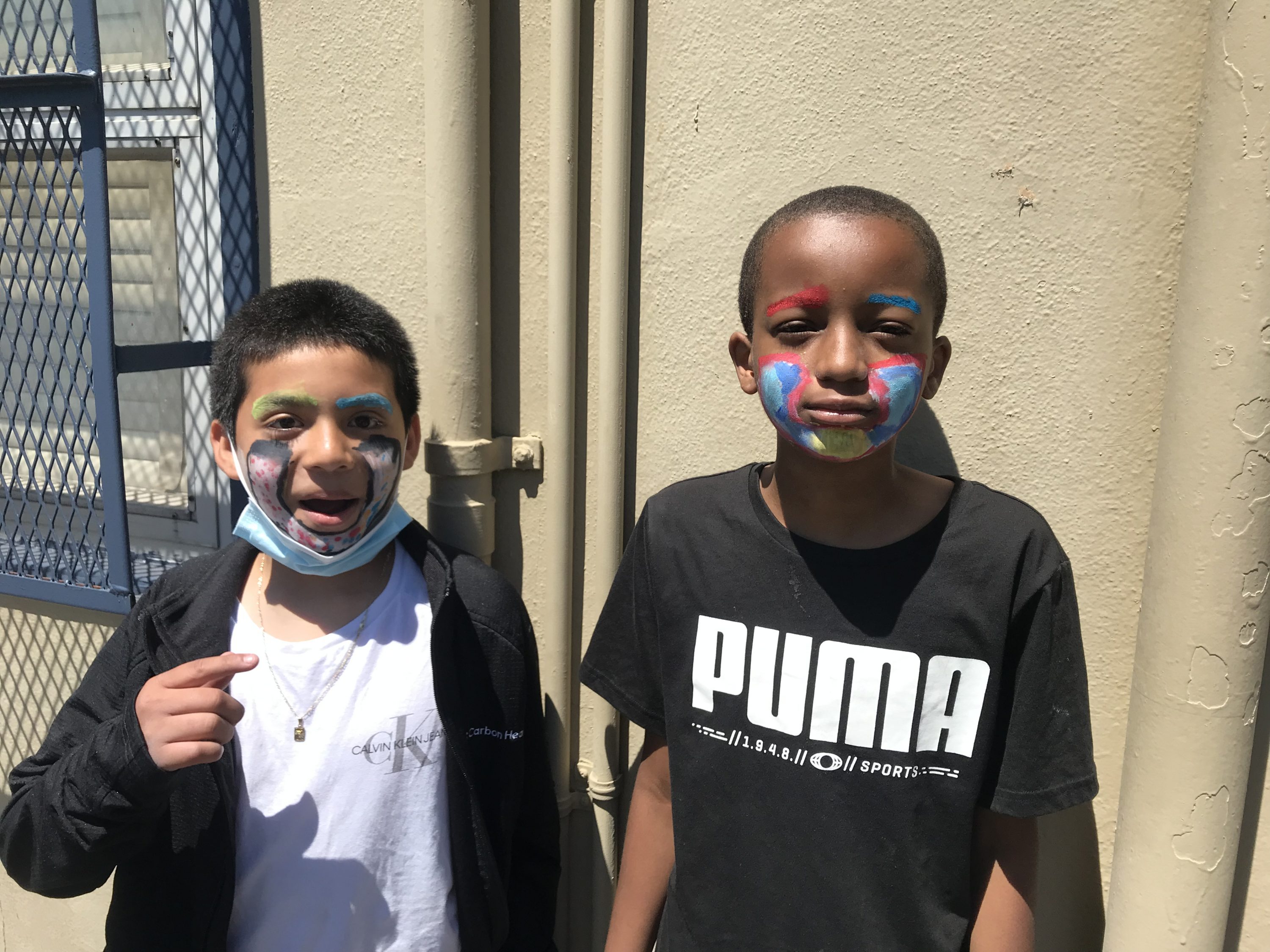 two children with brown and dark brown skin with colorful make up on their cheeks one is wearing a white t-shirt with a black jacket and the other is wearing a black t-shirt with the word "PUMA" on the chest