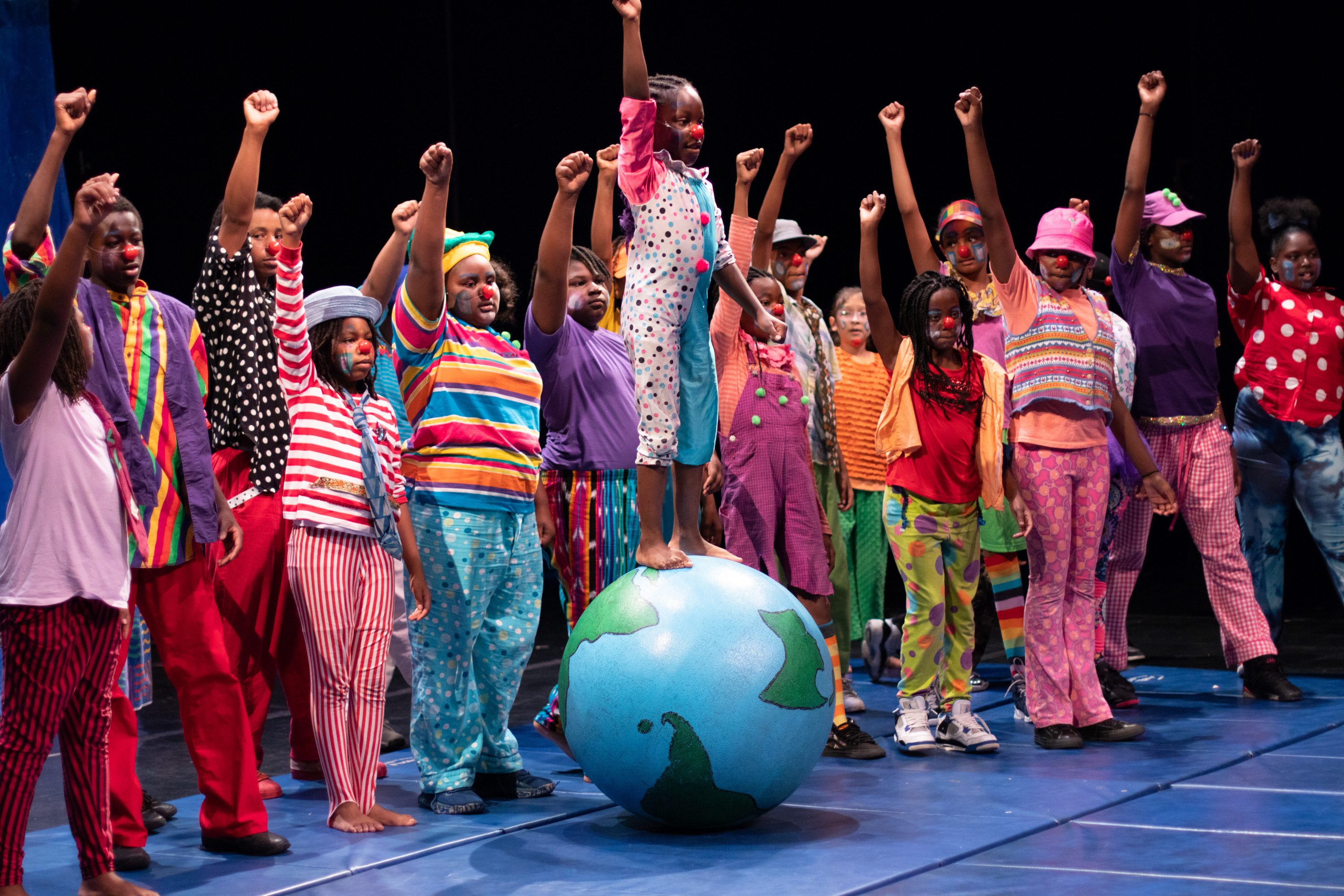 A young PCT performer dressed in a colorful costume is standing on top of a ball painted like the world with their right fist raised up. They are surrounded on each side by many other youth performers dressed in colorful costumes with raised fists. 
